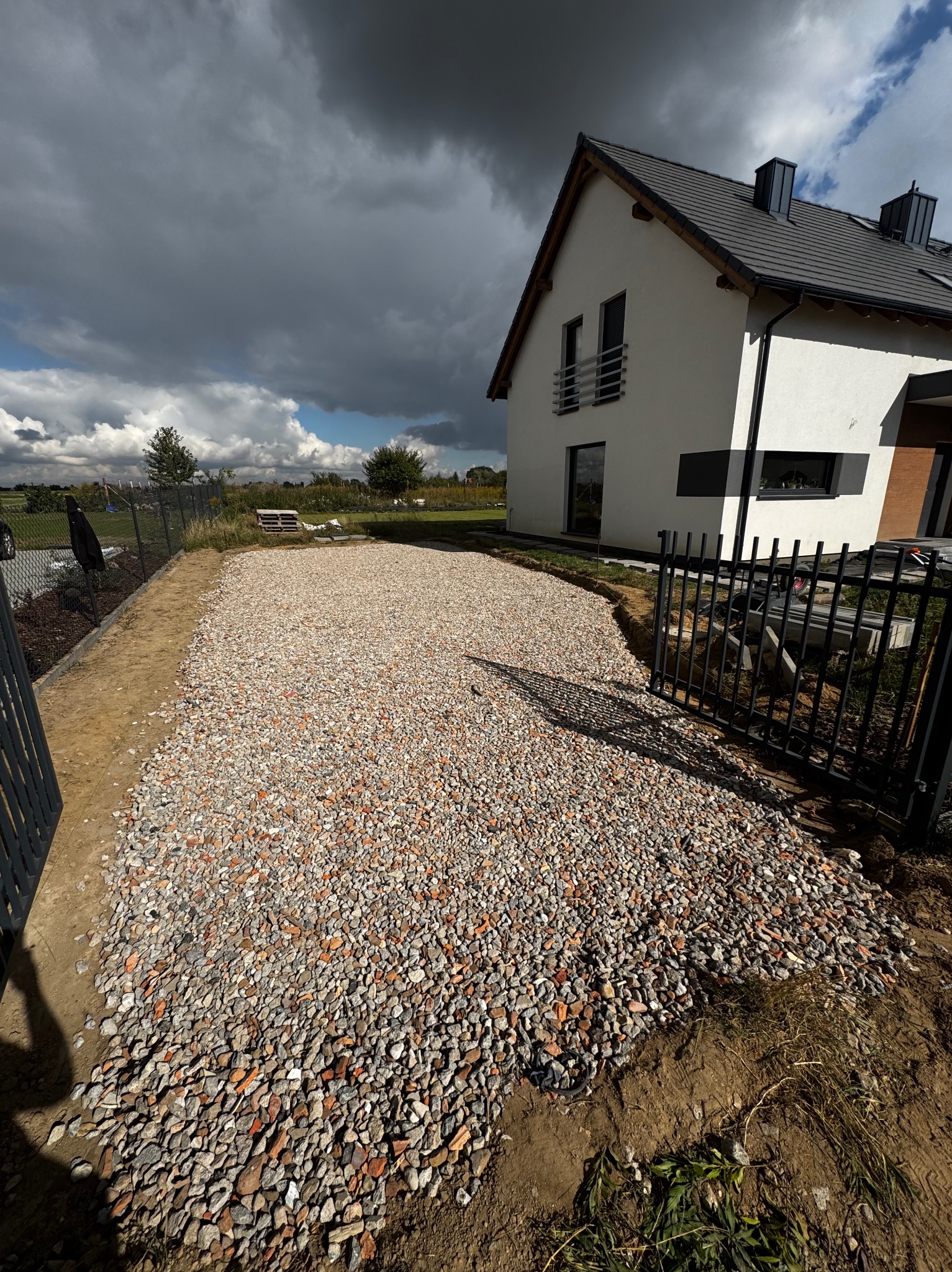 Teren przygotowany pod kostkę brukową z widoczną warstwą kruszywa, ogrodzeniem i domem w tle, pod pochmurnym niebem.