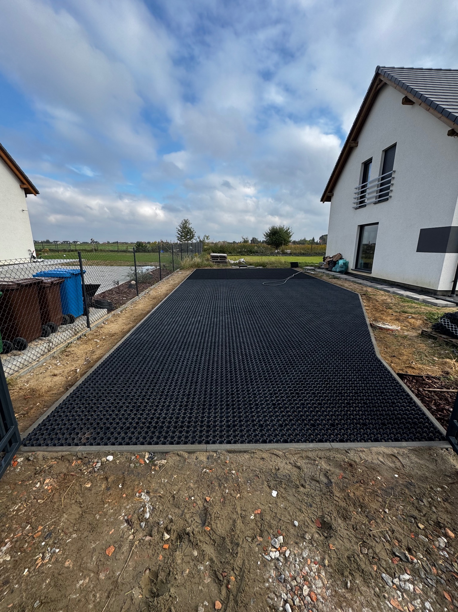 Czarna, ażurowa nawierzchnia na podwórku, obramowana szarym betonem, przygotowana pod kostkę. W tle dom i błękitne niebo z chmurami. Widok z poziomu gruntu.