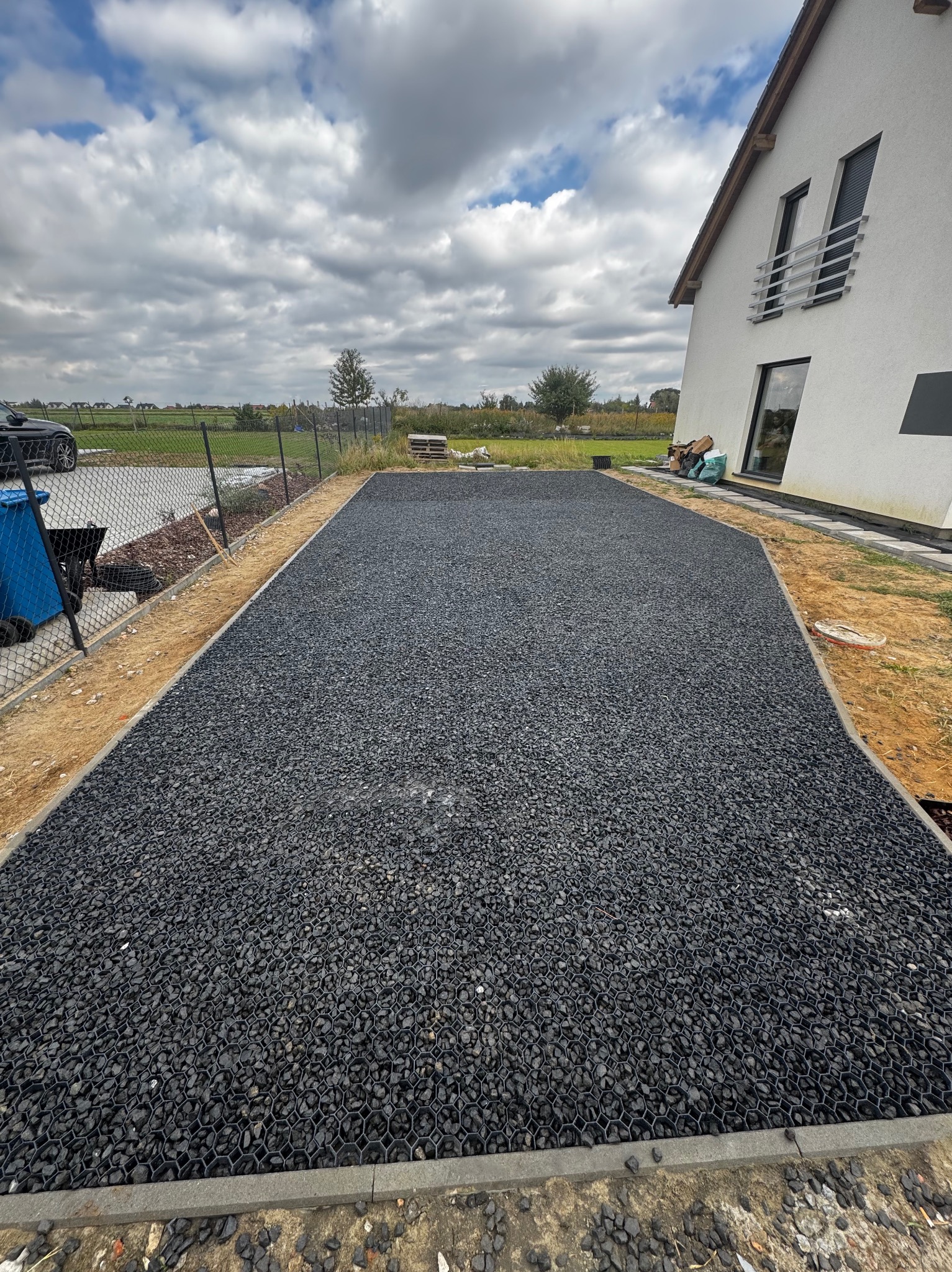 Ukończona powierzchnia z czarnego kruszywa wypełniającego plastikowe kratki, obrzeża betonowe. Dom z białym tynkiem w tle, pochmurne niebo nad posesją.