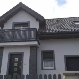 Nowoczesny dom jednorodzinny z szarą elewacją, ciemnoszarą stolarką okienną i balkonem, oraz grafitowym dachem z dachówki ceramicznej.