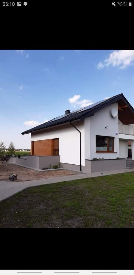 Nowoczesny dom jednorodzinny z białą elewacją, drewnianymi akcentami i szarą podmurówką, widok z poziomu trawnika w słoneczny dzień.