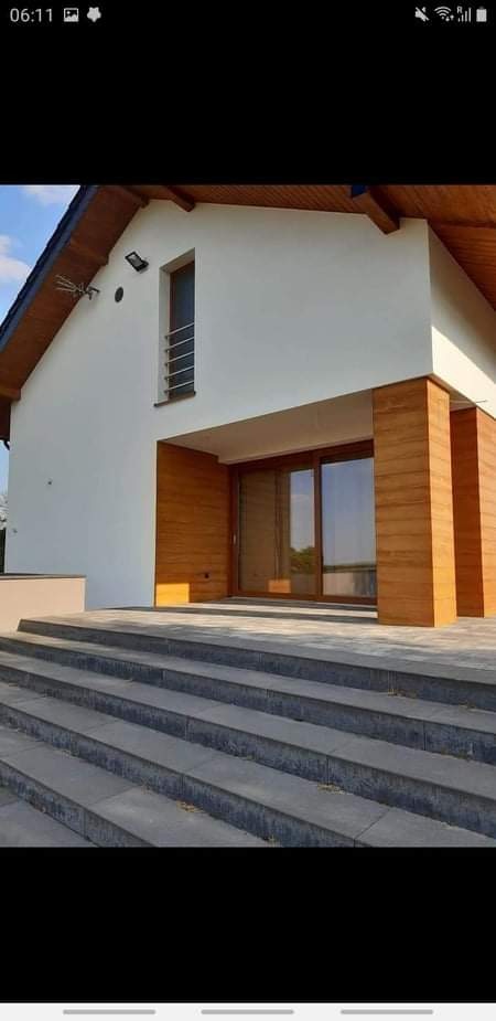 Nowoczesny dom z białymi ścianami, drewnianą okładziną przy wejściu i schodami z szarego kamienia prowadzącymi do drzwi balkonowych.