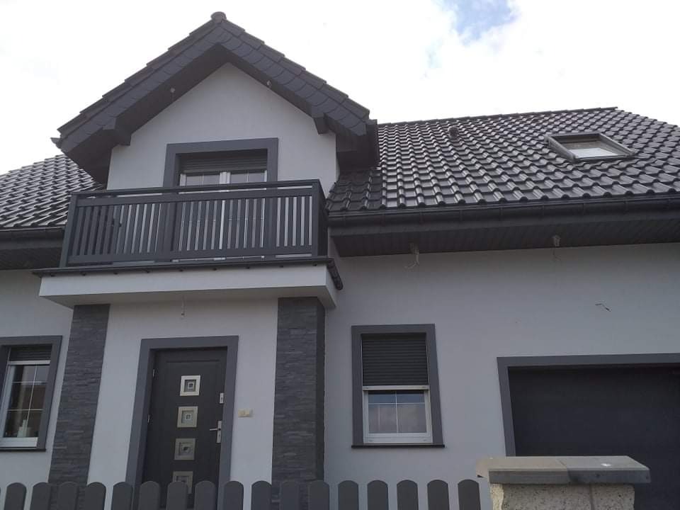 Nowoczesny dom jednorodzinny z szarą elewacją, ciemnoszarą stolarką okienną i balkonem, oraz grafitowym dachem z dachówki ceramicznej.