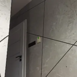 Szara ściana z dekoracyjną fakturą i czarnymi liniami tworzącymi geometryczny wzór, obok białe drzwi z czarną klamką, widoczny fragment sufitu i podłogi.