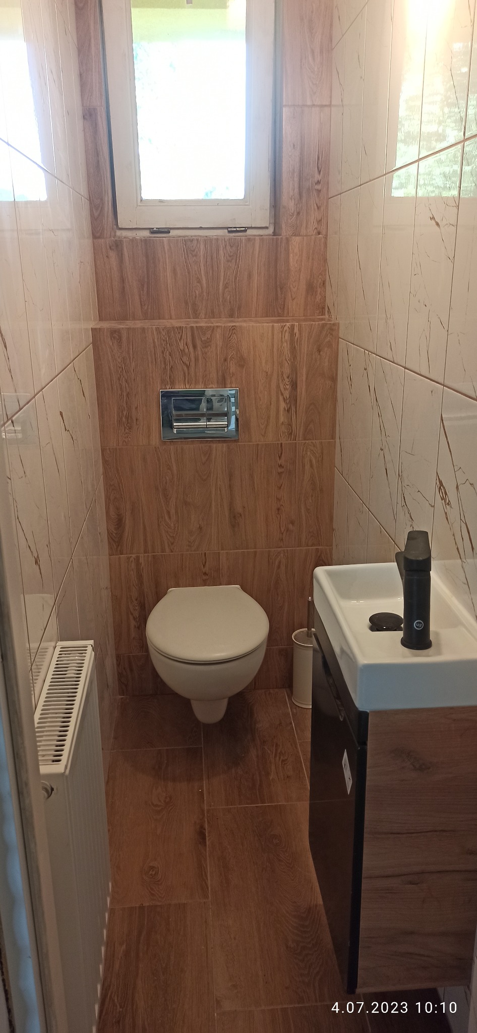 Wąska łazienka po remoncie, z brązowymi płytkami imitującymi drewno na ścianach i podłodze, podwieszana toaleta, biała umywalka z czarną baterią i szafką pod umywalką, grzejnik drabinkowy po lewej...