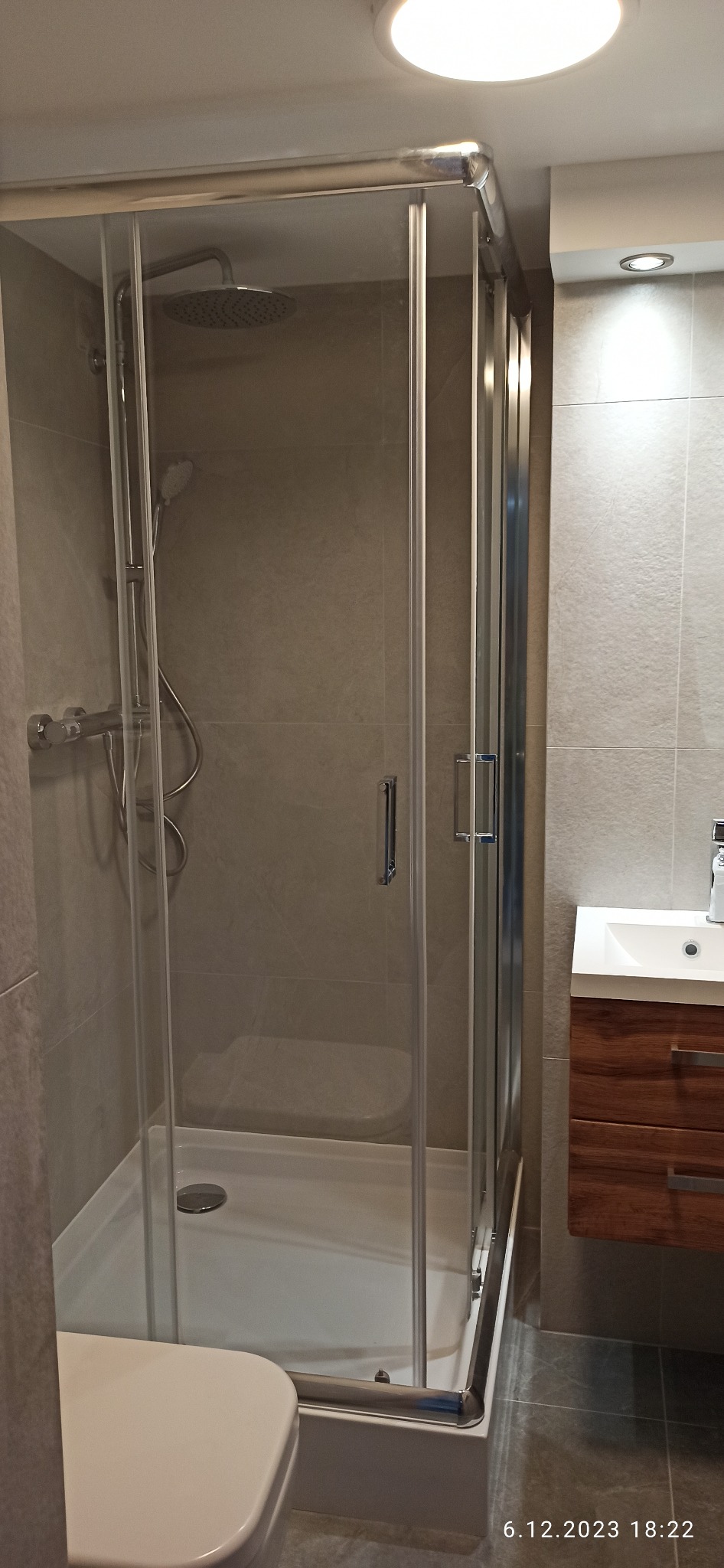 Nowoczesna kabina prysznicowa z deszczownicą, szklane drzwi z chromowanymi uchwytami, szare płytki na ścianach, umywalka z drewnianą szafką w tle.