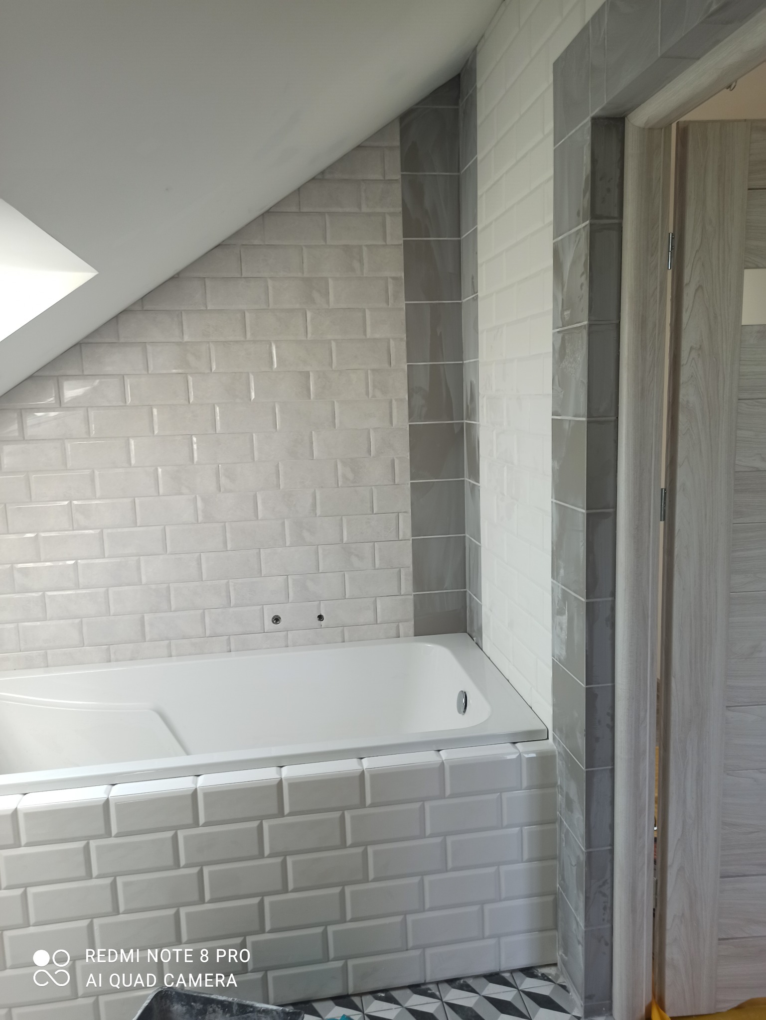 Wykończona łazienka na poddaszu z białą wanną obudowaną kafelkami typu metro, szare płytki akcentujące narożniki i wejście, oraz wzorzystą posadzką.