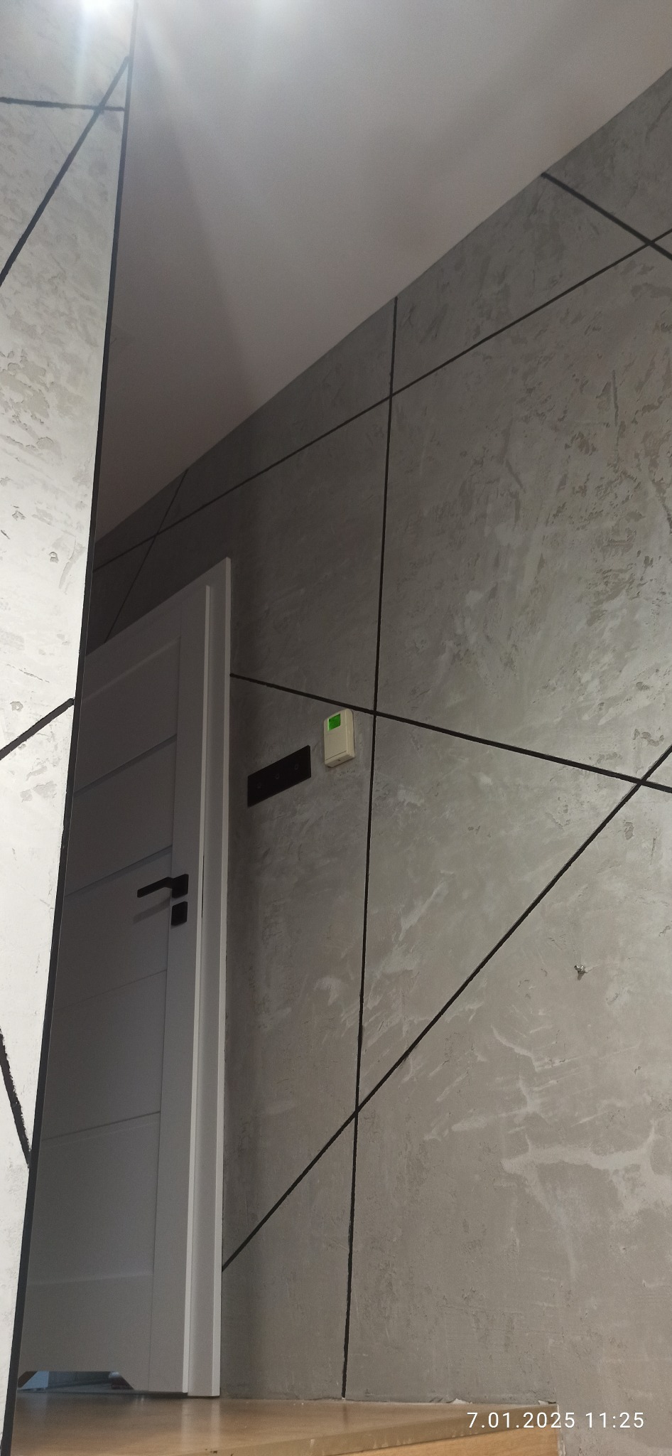 Szara ściana z dekoracyjną fakturą i czarnymi liniami tworzącymi geometryczny wzór, obok białe drzwi z czarną klamką, widoczny fragment sufitu i podłogi.