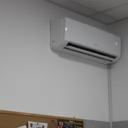 Biały klimatyzator ścienny z wyświetlaczem temperatury 30 stopni, zamontowany w biurze nad tablicą korkową i listwą przypodłogową z efektem lastryko.