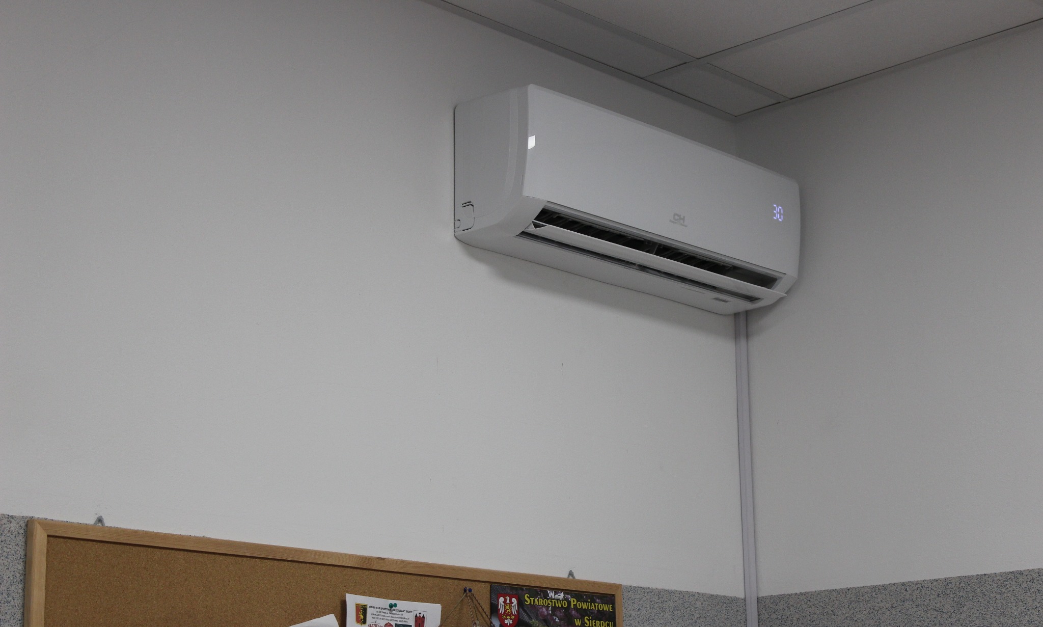 Biały klimatyzator ścienny z wyświetlaczem temperatury 30 stopni, zamontowany w biurze nad tablicą korkową i listwą przypodłogową z efektem lastryko.