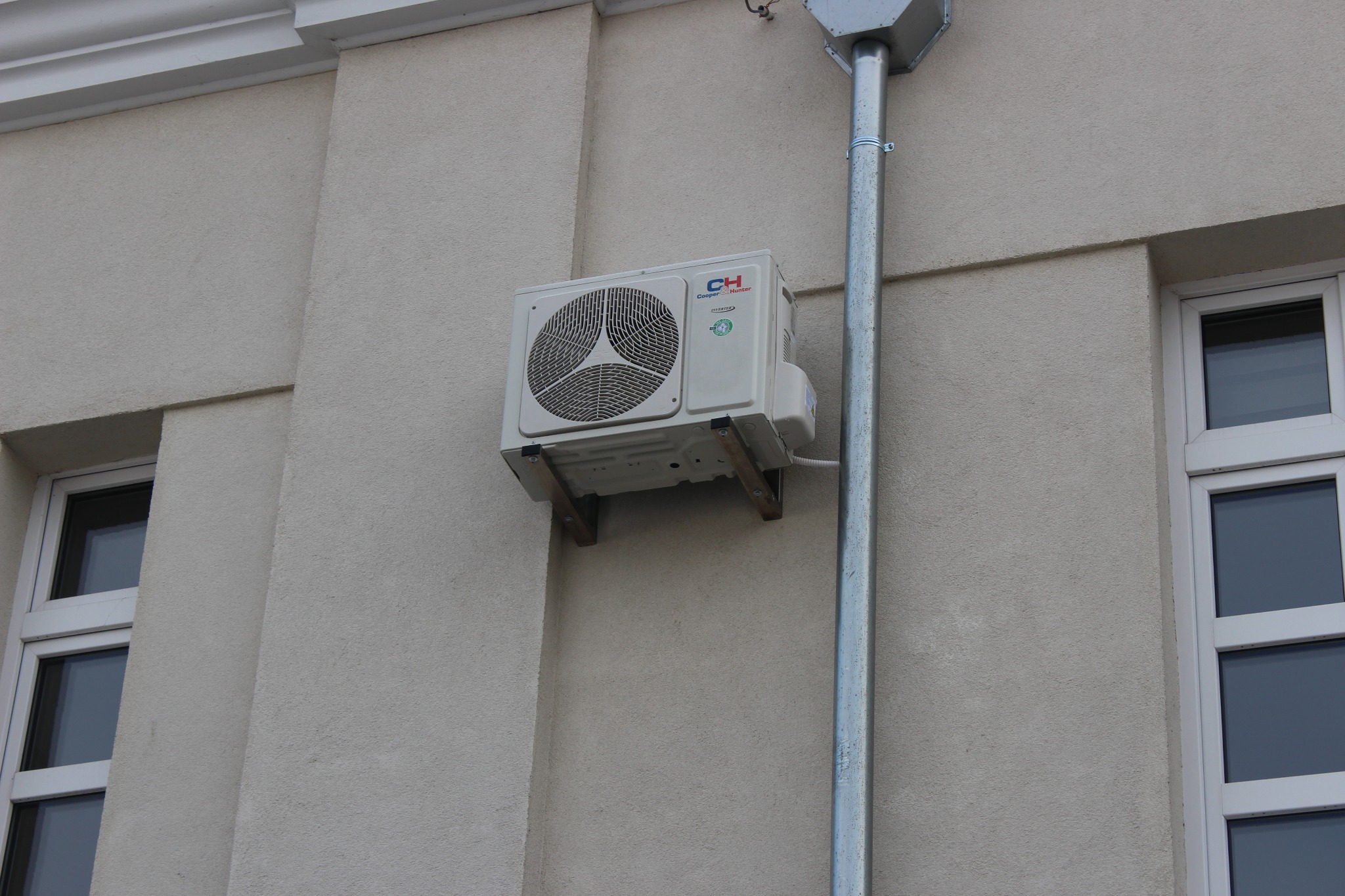Biały klimatyzator Cooper&Hunter zamontowany na zewnątrz budynku na metalowych wspornikach, obok rynny spustowej.
