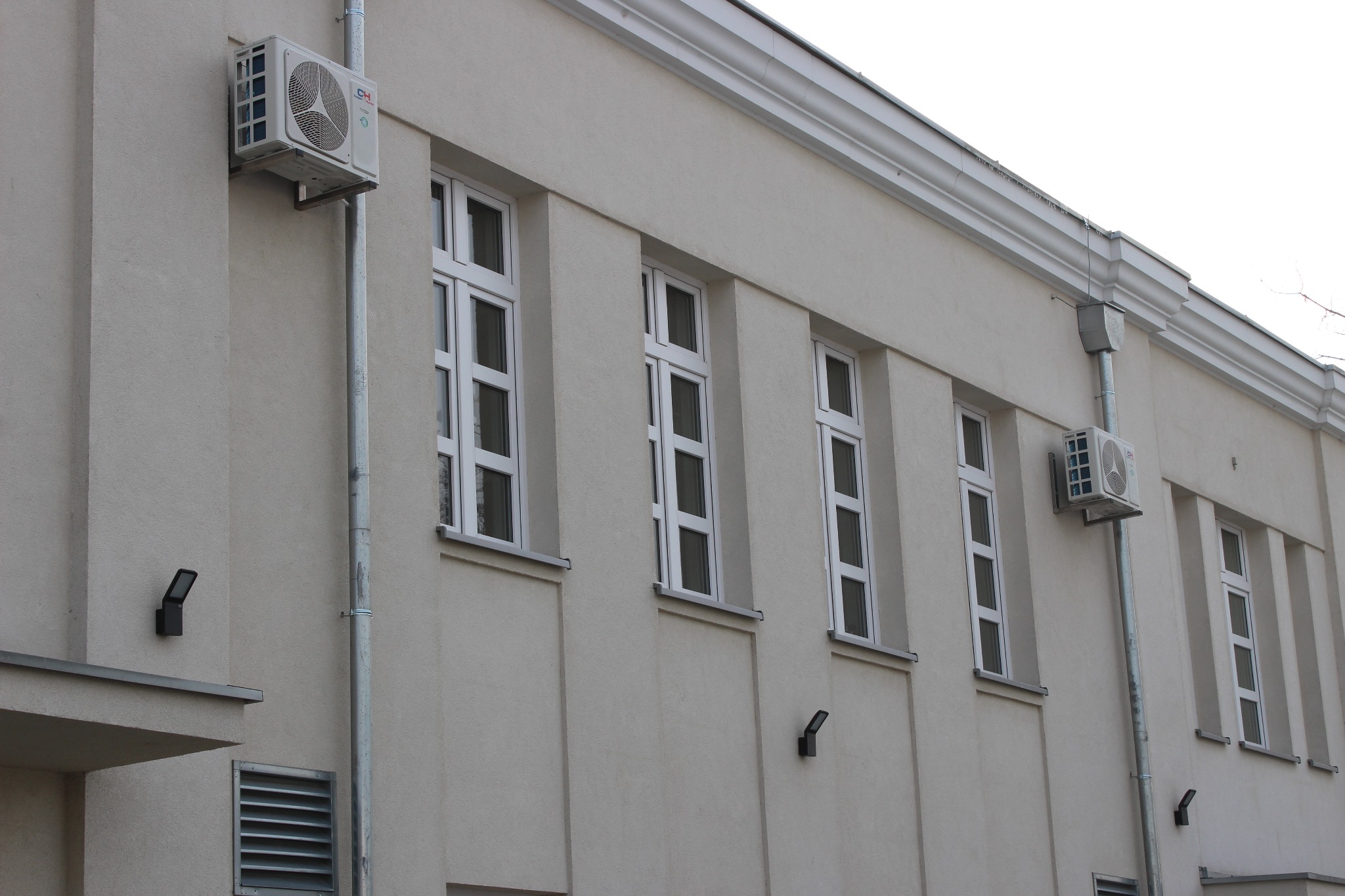 Elewacja budynku z zamontowanymi jednostkami zewnętrznymi klimatyzacji, rynnami i oknami z białymi ramami.