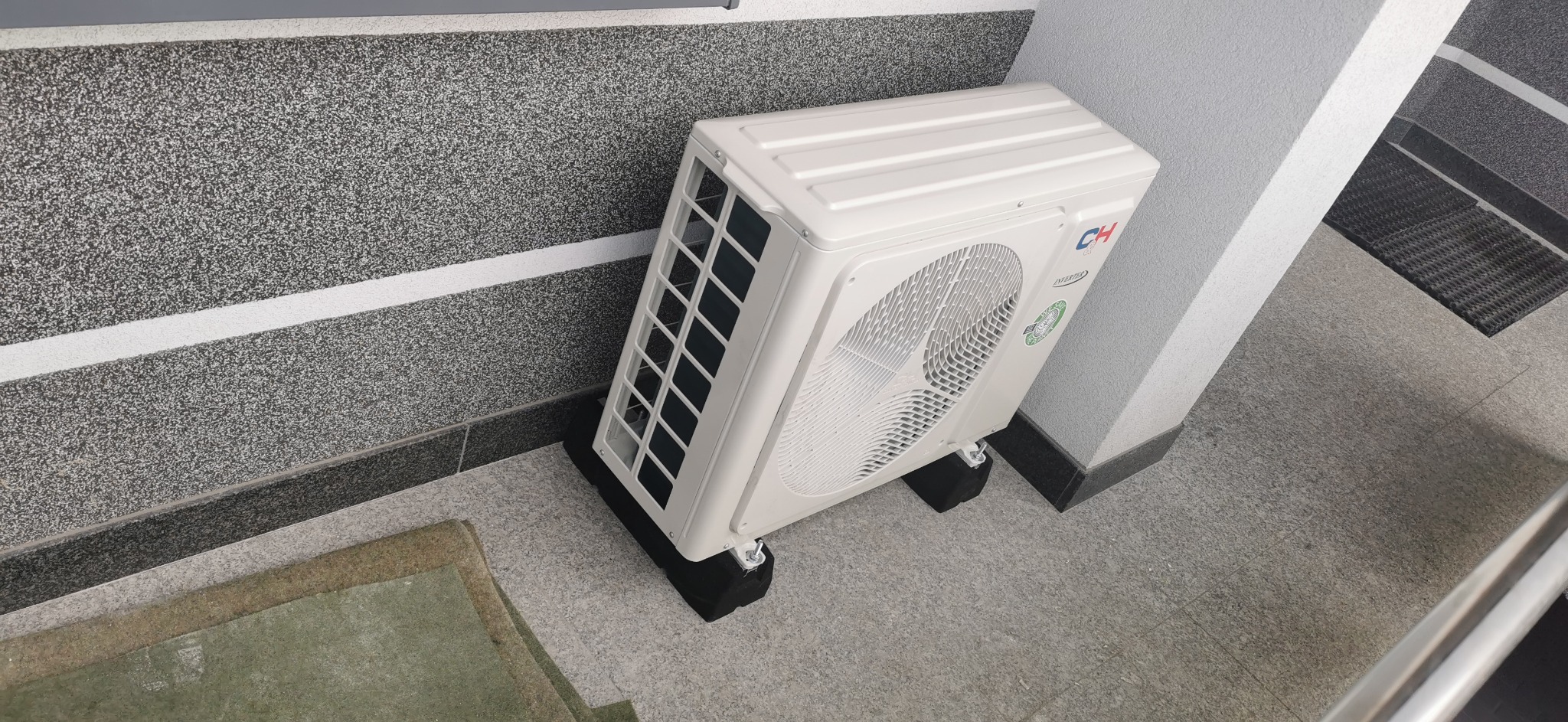 Biała jednostka zewnętrzna klimatyzacji marki CH zamontowana na czarnych podkładkach na szarych płytkach podłogowych, widok z góry, ściana w tle z szarego lastryko.