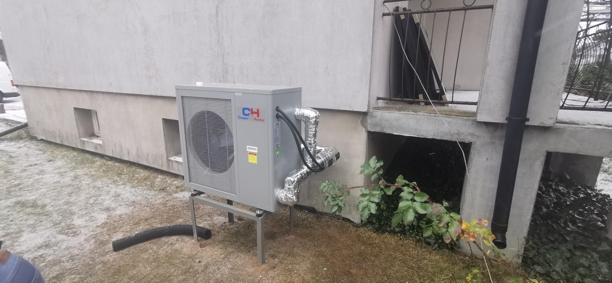 Zewnętrzna jednostka pompy ciepła Cooper&Hunter zamontowana na metalowym stelażu przy ścianie budynku z widocznymi przewodami w srebrnej izolacji oraz fragmentem czarnej rury na ziemi.