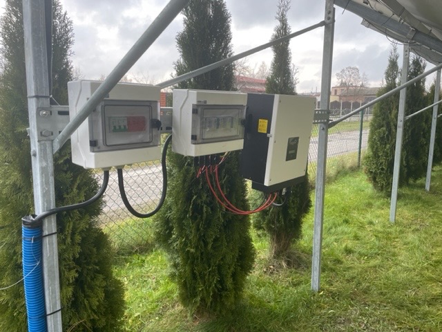 Dwa białe panele rozdzielcze i falownik zamontowane na metalowej konstrukcji przy tujach, widoczne kable i rurka karbowana.