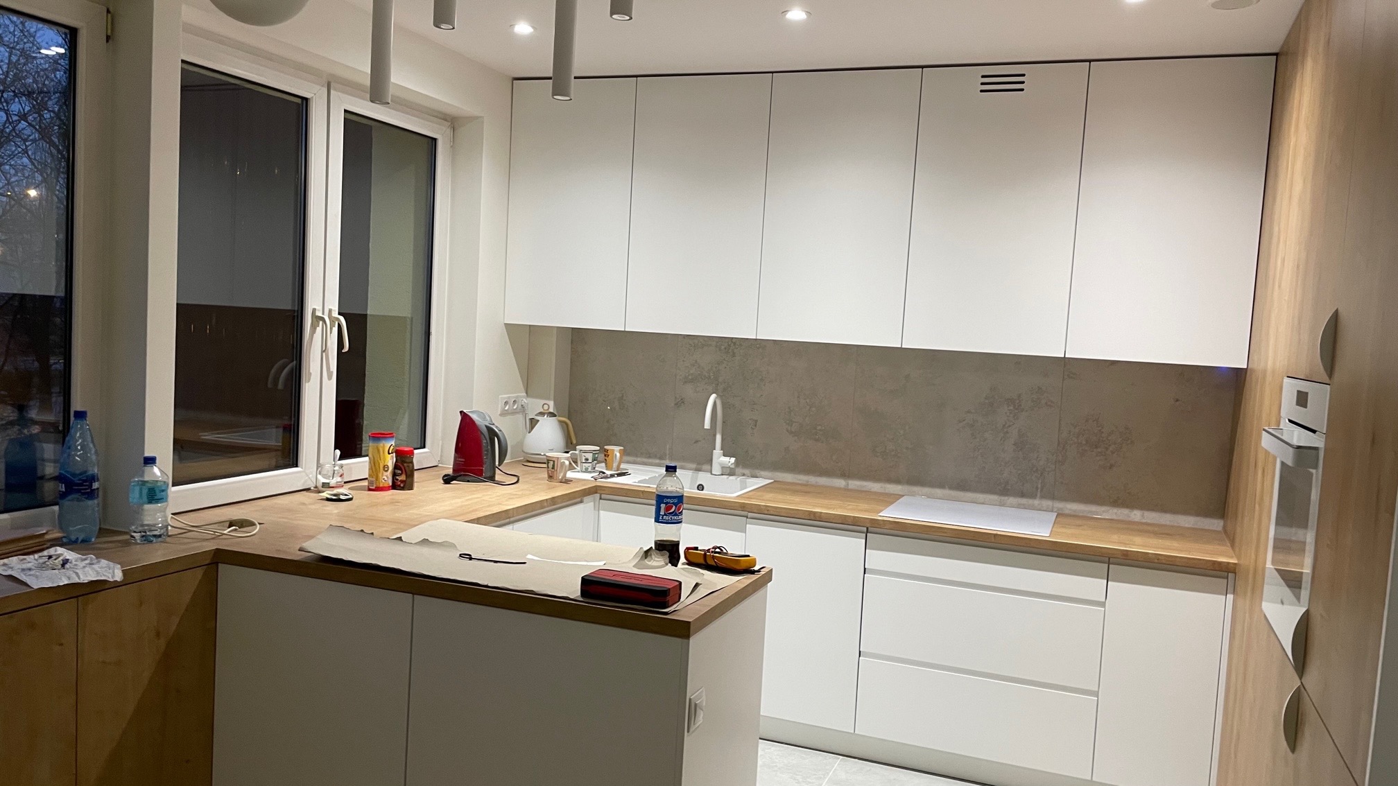 Nowoczesna kuchnia z białymi szafkami i drewnianym blatem, wyposażona w sprzęt AGD, z widocznymi śladami prac wykończeniowych na blacie.