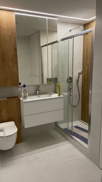 Nowoczesna łazienka z wiszącą szafką umywalkową, kabiną prysznicową ze szklanymi drzwiami przesuwnymi i drewnianymi akcentami na ścianie obok prysznica. W tle lustro z oświetleniem LED.