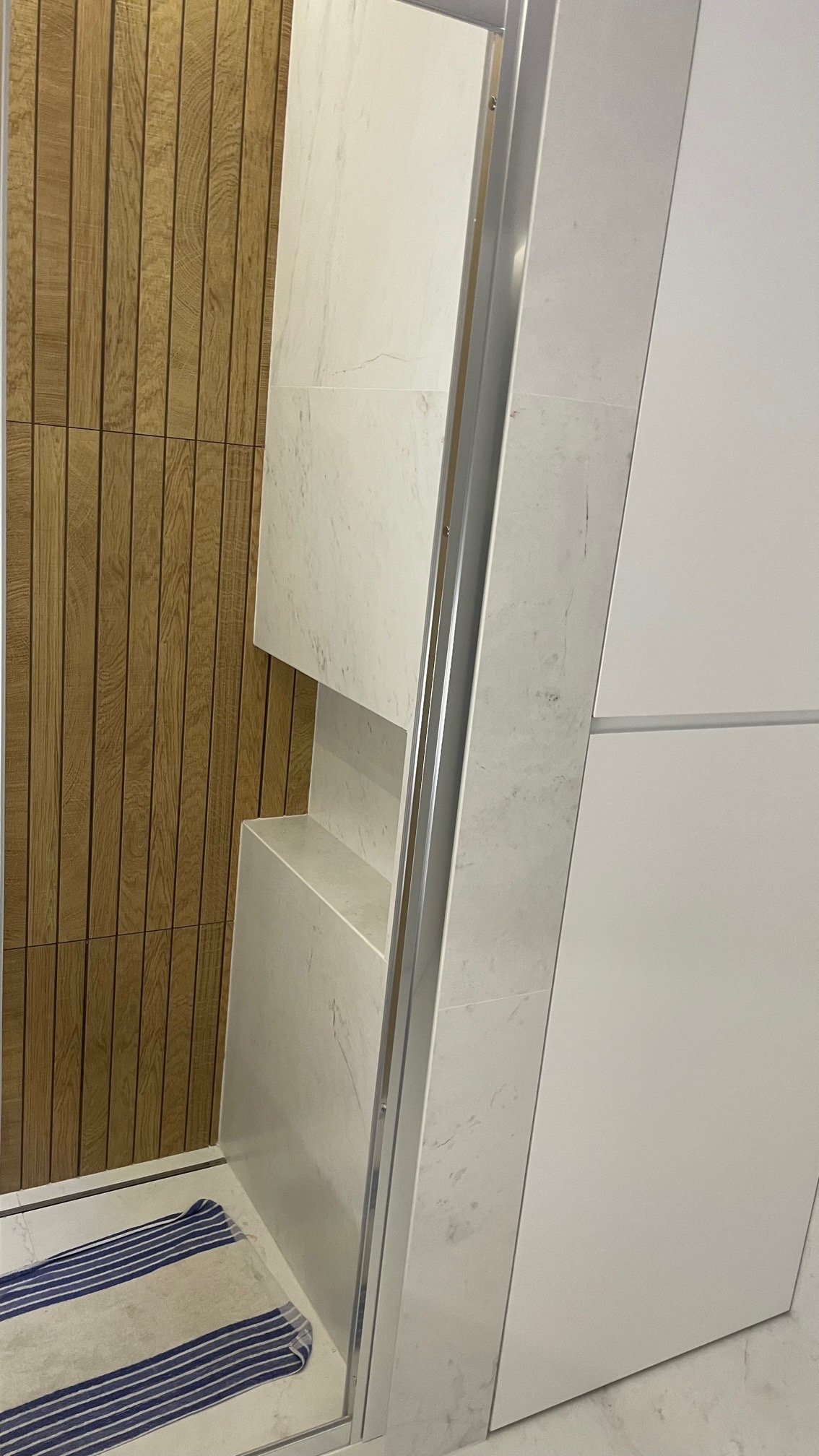 Nowoczesna łazienka z drewnianą ścianą, kabiną prysznicową z marmurowymi elementami i wnękami oraz ręcznikiem w paski na podłodze.