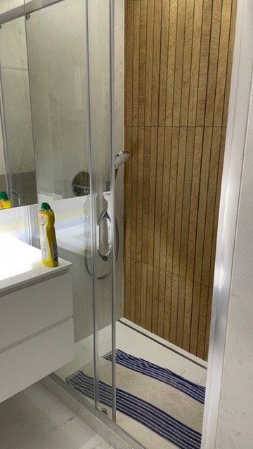 Kabina prysznicowa z przesuwnymi drzwiami, wyłożona drewnopodobnymi panelami pionowymi, widoczny fragment umywalki z płynem do czyszczenia i niebiesko-biały chodnik wewnątrz.