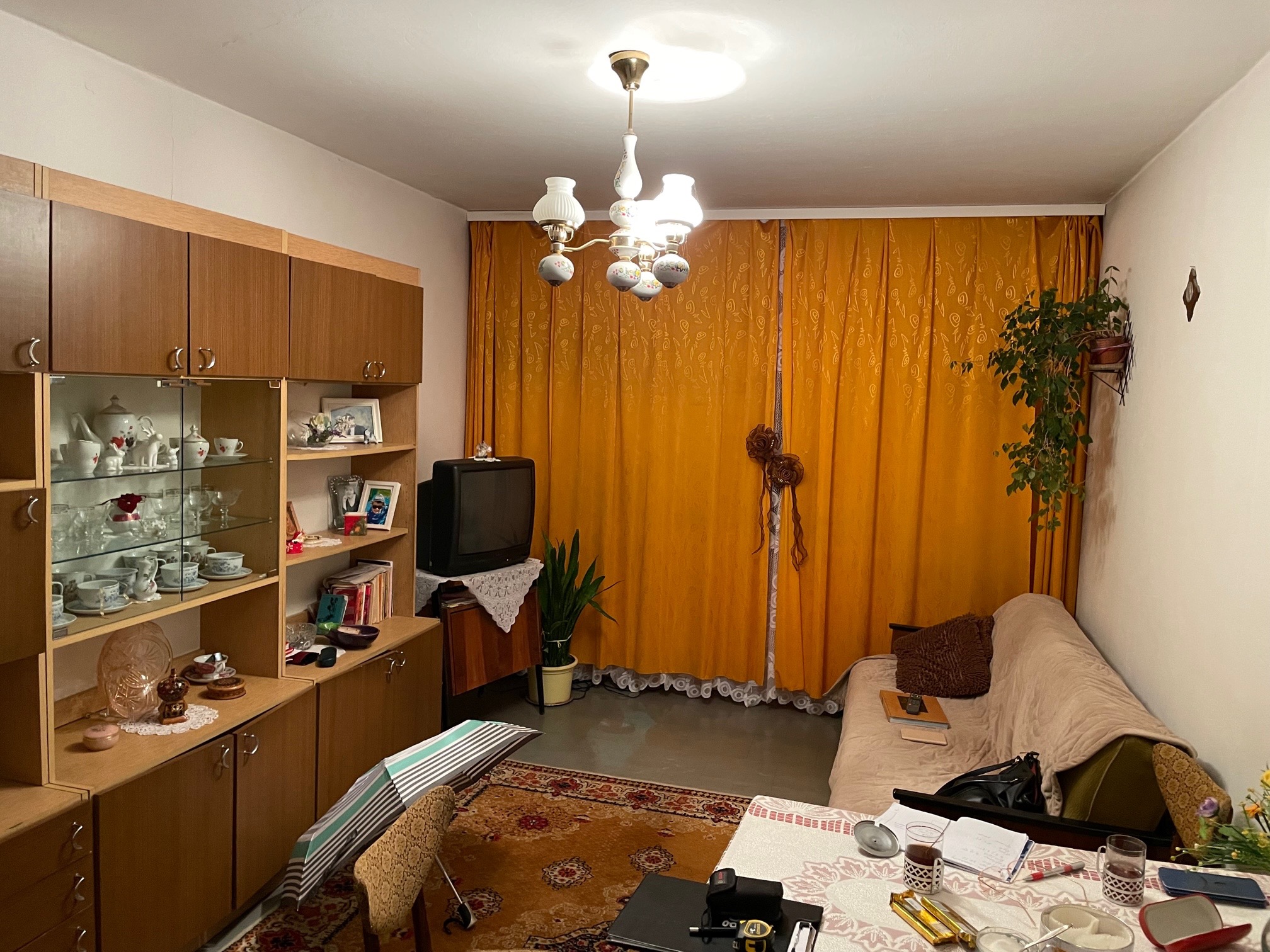 Pokój dzienny z meblościanką, zasłonami w kolorze musztardowym, dywanem w orientalne wzory i lampą sufitową w stylu retro. Na stole widoczne przedmioty codziennego użytku.