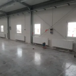 Wnętrze hali przemysłowej z zamontowanymi, białymi grzejnikami pod oknami. Na podłodze widoczny worek i zielone wiadro.