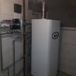 Biały piec centralnego ogrzewania z podłączonymi rurami miedzianymi i stalowymi w kotłowni wyłożonej szarymi płytkami.