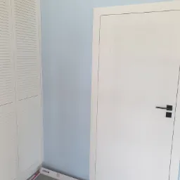 Fragment pokoju w trakcie prac wykończeniowych: biała szafa z żaluzjowymi drzwiami, błękitna ściana, białe drzwi z czarną klamką oraz panele podłogowe w opakowaniach przygotowane do ułożenia.