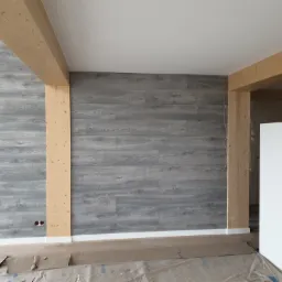 Ściana wykończona panelami imitującymi drewno w odcieniach szarości, z widocznymi drewnianymi belkami konstrukcyjnymi i zabezpieczoną podłogą podczas prac wykończeniowych.