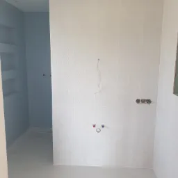 Jasna łazienka w trakcie wykańczania, widoczne świeżo położone białe płytki z delikatnym wzorem jodełki na ścianach, podłoga również wyłożona jasnymi płytkami, wystające kable i rury wodne...