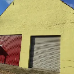 Żółty budynek z dwoma bramami garażowymi: czerwoną pionową i szarą rolowaną. Widoczne ślady po pnączach na ścianie.