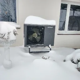 Szara pompa ciepła marki Nesheat, częściowo przysypana śniegiem, stoi na zewnątrz budynku w zimowej scenerii.