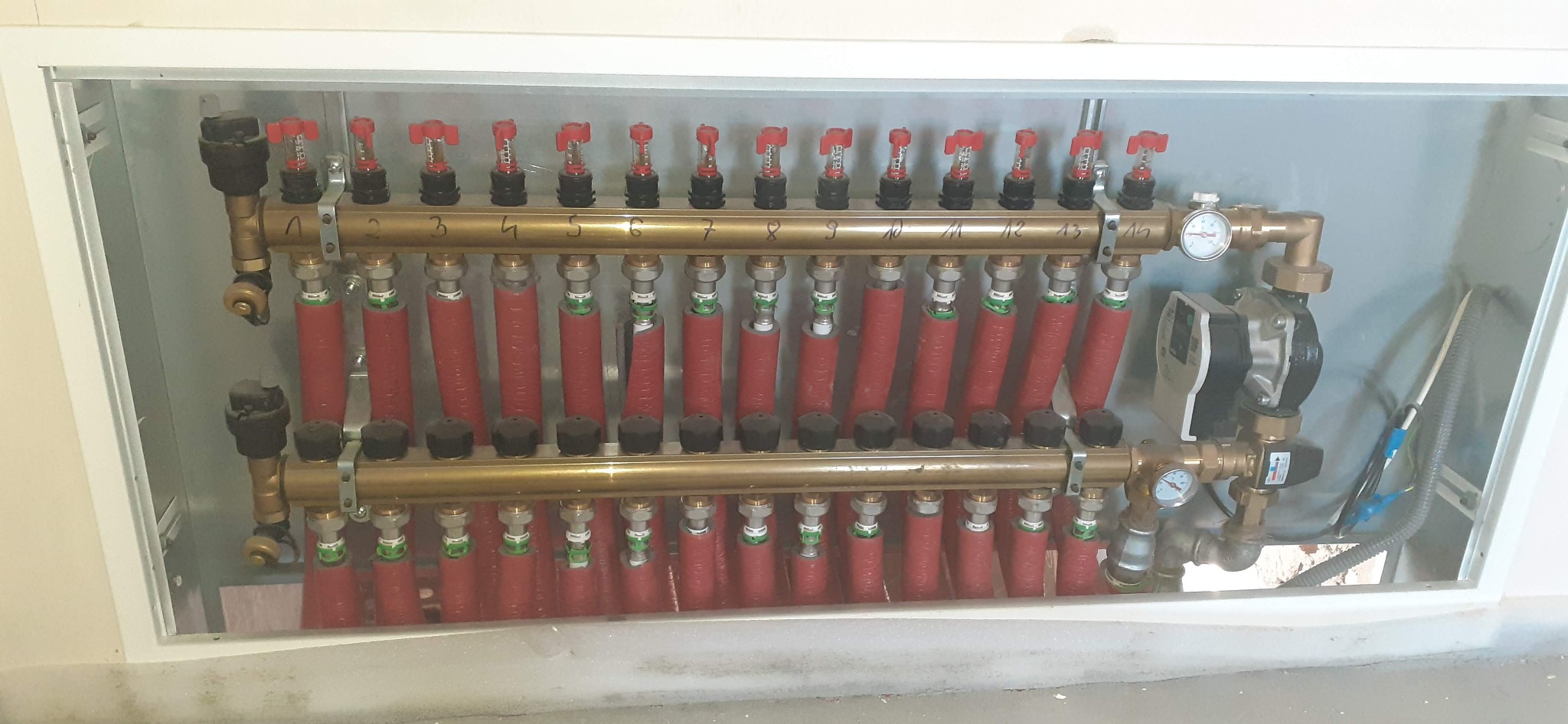 Rozdzielacz ogrzewania podłogowego z czternastoma obwodami, widoczne rury w izolacji termicznej, manometry i pompa obiegowa w białej szafce instalacyjnej.