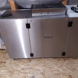 Srebrny rekuperator marki reQ-net z podłączonymi przewodami wentylacyjnymi, umieszczony na drewnianej podłodze na poddaszu.