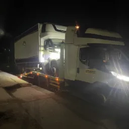 Nocne ujęcie lawety Iveco z załadowanym busem. Widoczna tablica z numerem telefonu pomocy drogowej. Oświetlenie uliczne i reflektory lawety.