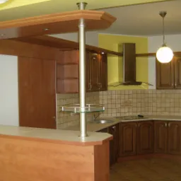 Wykończona kuchnia z drewnianymi szafkami w klasycznym stylu, wyspą z blatem i słupem z półką, oraz okapem nad płytą grzewczą.