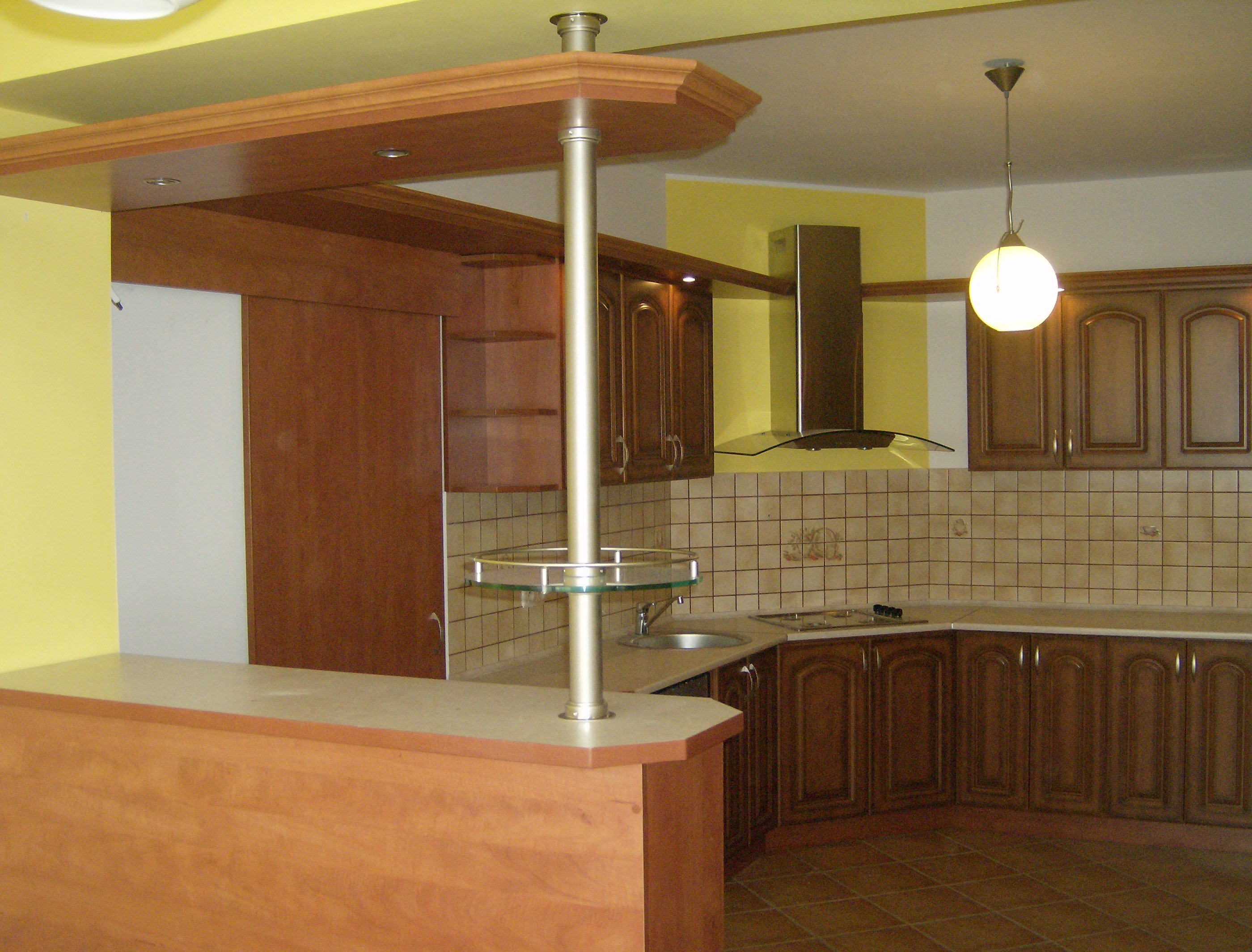 Wykończona kuchnia z drewnianymi szafkami w klasycznym stylu, wyspą z blatem i słupem z półką, oraz okapem nad płytą grzewczą.