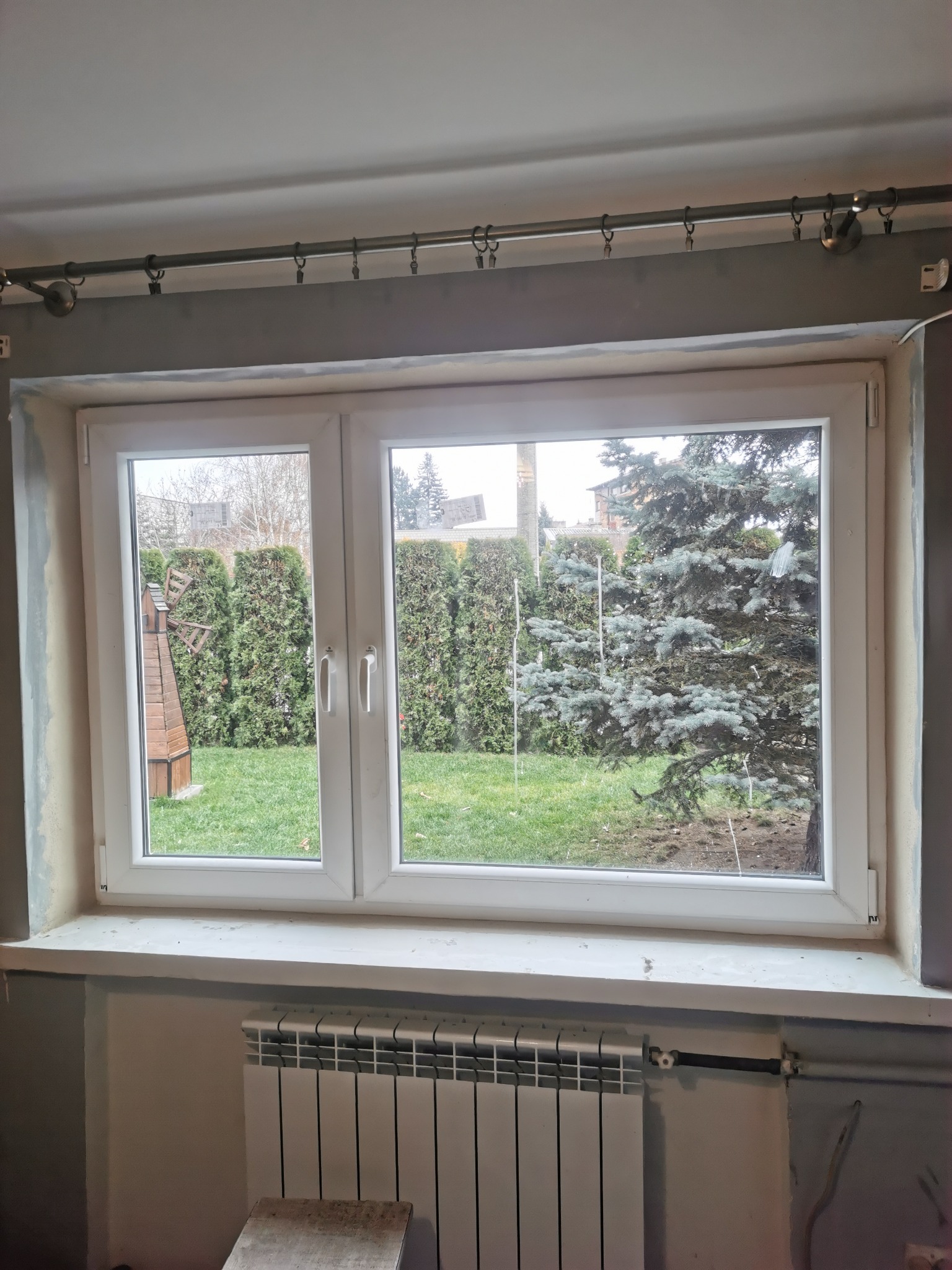 Dwuskrzydłowe okno PCV z widokiem na ogród z drewnianym wiatrakiem i iglakiem, ściana z widocznym grzejnikiem i śladami po remoncie.