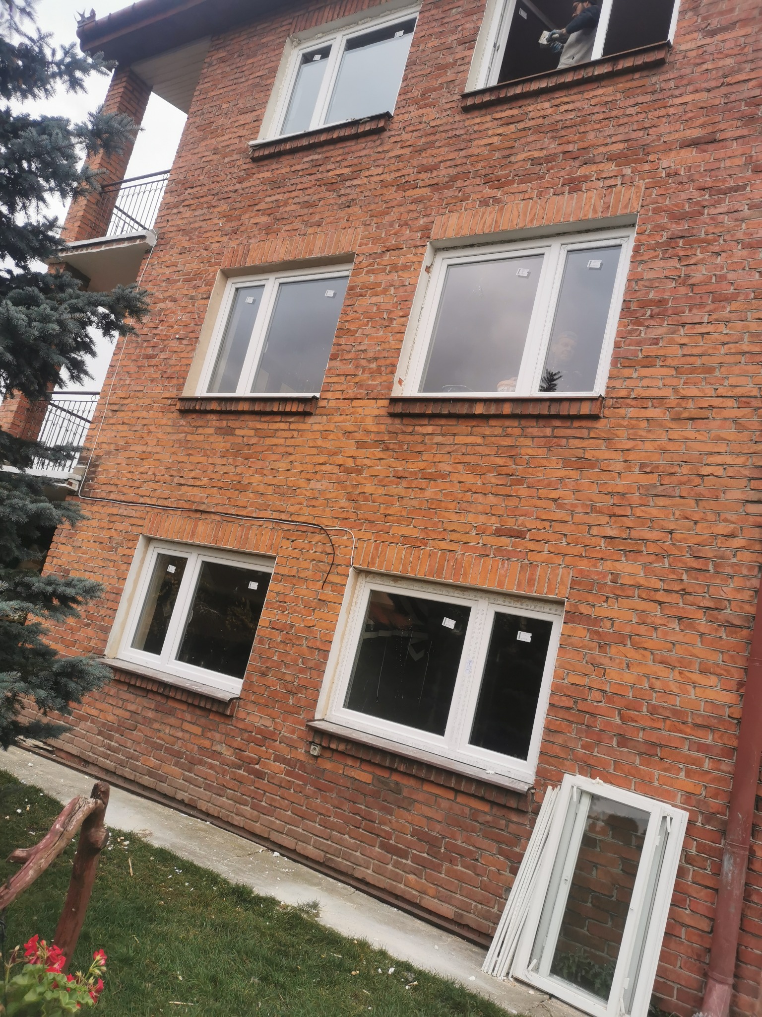 Dwupiętrowy budynek z czerwonej cegły z białymi oknami; na najwyższym piętrze widoczna osoba w kombinezonie roboczym, na trawniku leżą zdemontowane ramy okienne.