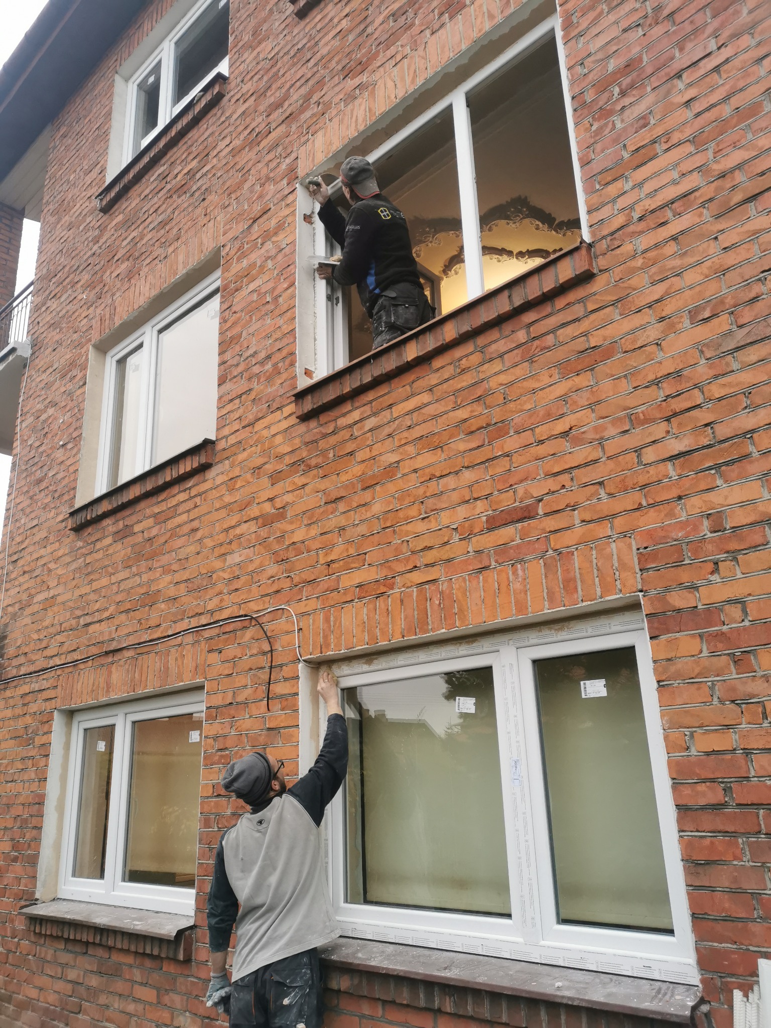 Dwóch monterów naprawia okna w bloku z czerwonej cegły, jeden uszczelnia okno na wysokości, drugi uszczelnia okno na parterze.
