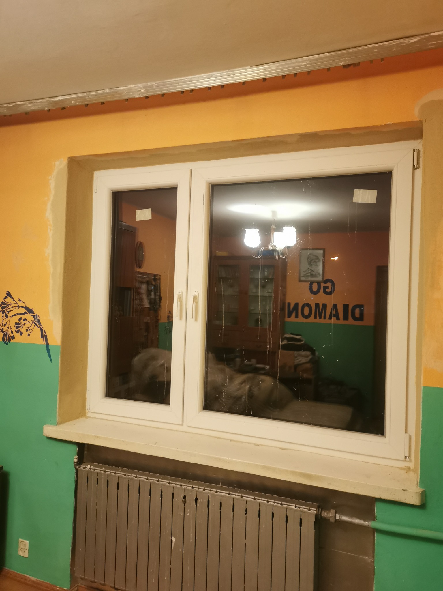 Białe plastikowe okno dwuskrzydłowe z widocznym grzejnikiem pod spodem, ściany pomalowane na żółto i zielono, widoczne ślady po remoncie.