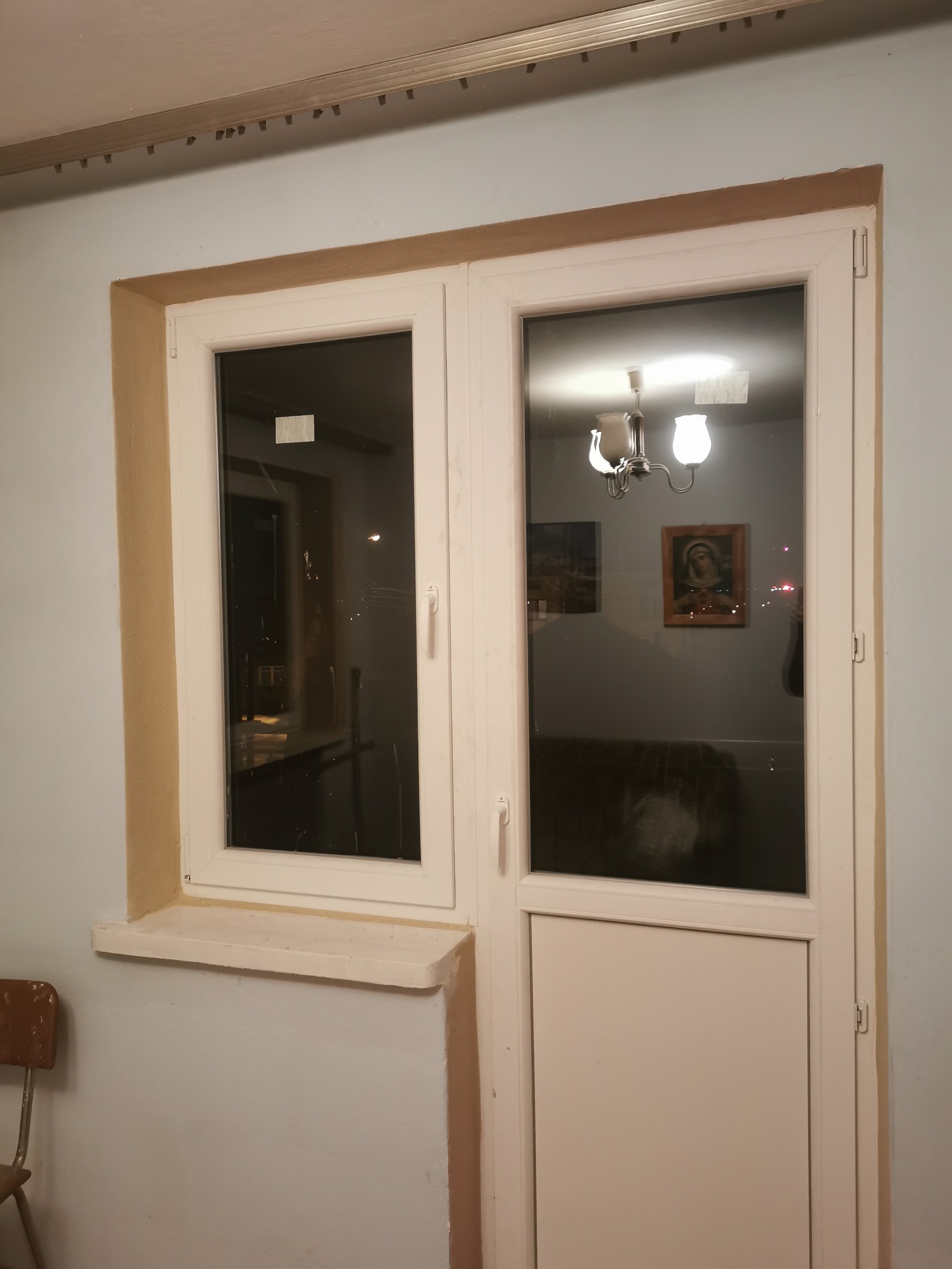 Białe okno PCV z parapetem, widoczne ślady niedokładnego malowania ramy okiennej i ściany, w szybie odbija się wnętrze z żyrandolem i obrazami.