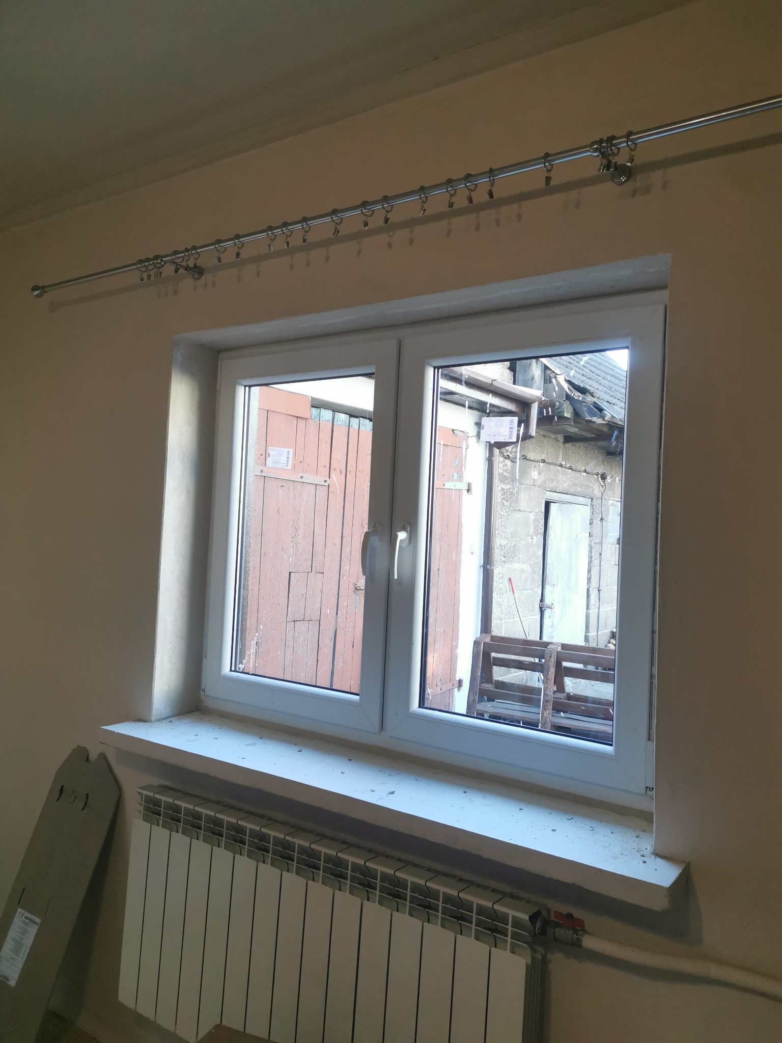 Białe okno PCV z widokiem na podwórze, zainstalowane w pokoju z grzejnikiem i karniszem, widoczne ślady po remoncie.