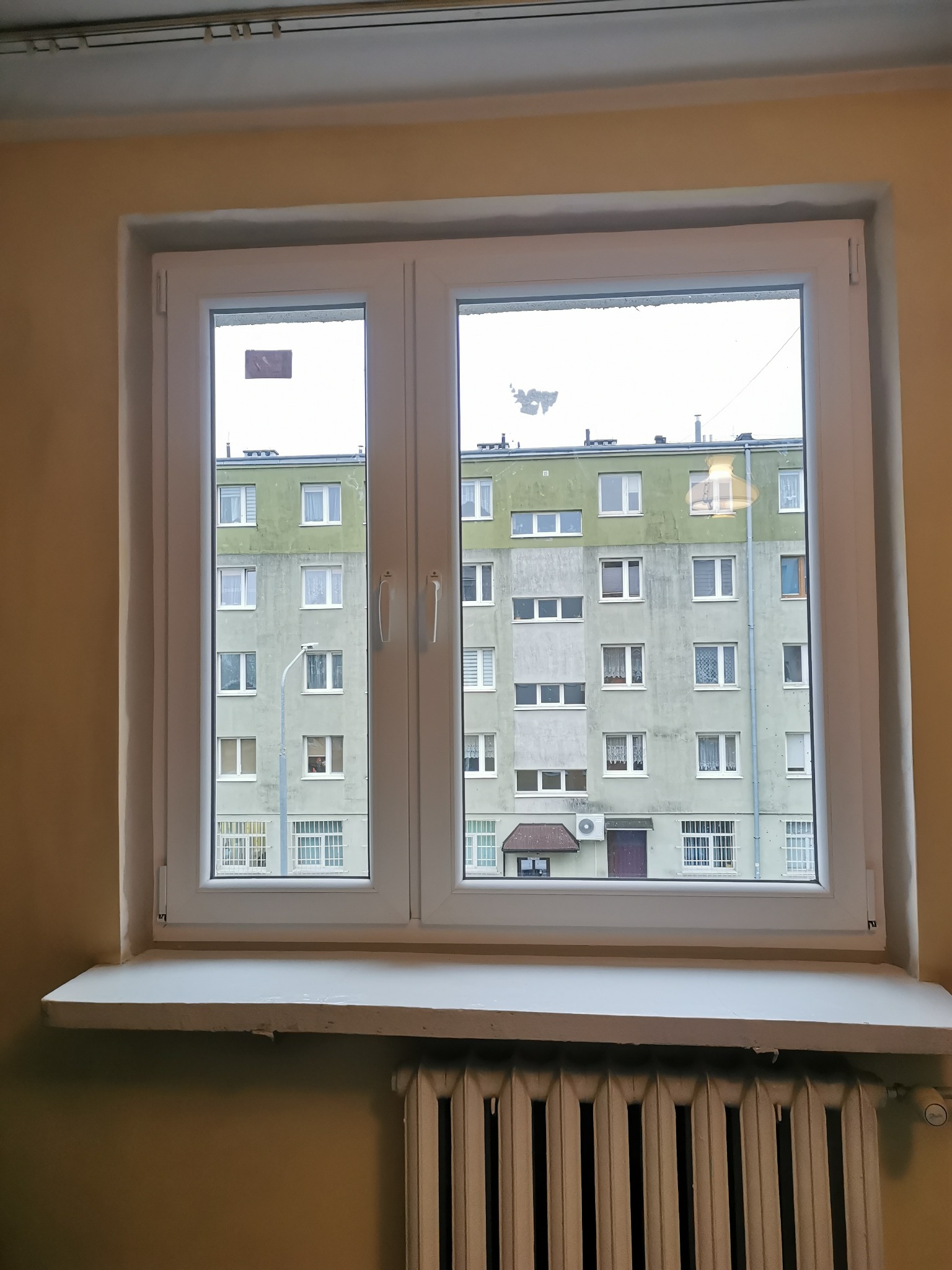 Białe, dwuskrzydłowe okno PCV z widokiem na blok mieszkalny, grzejnik pod parapetem, ściana w odcieniu beżu.