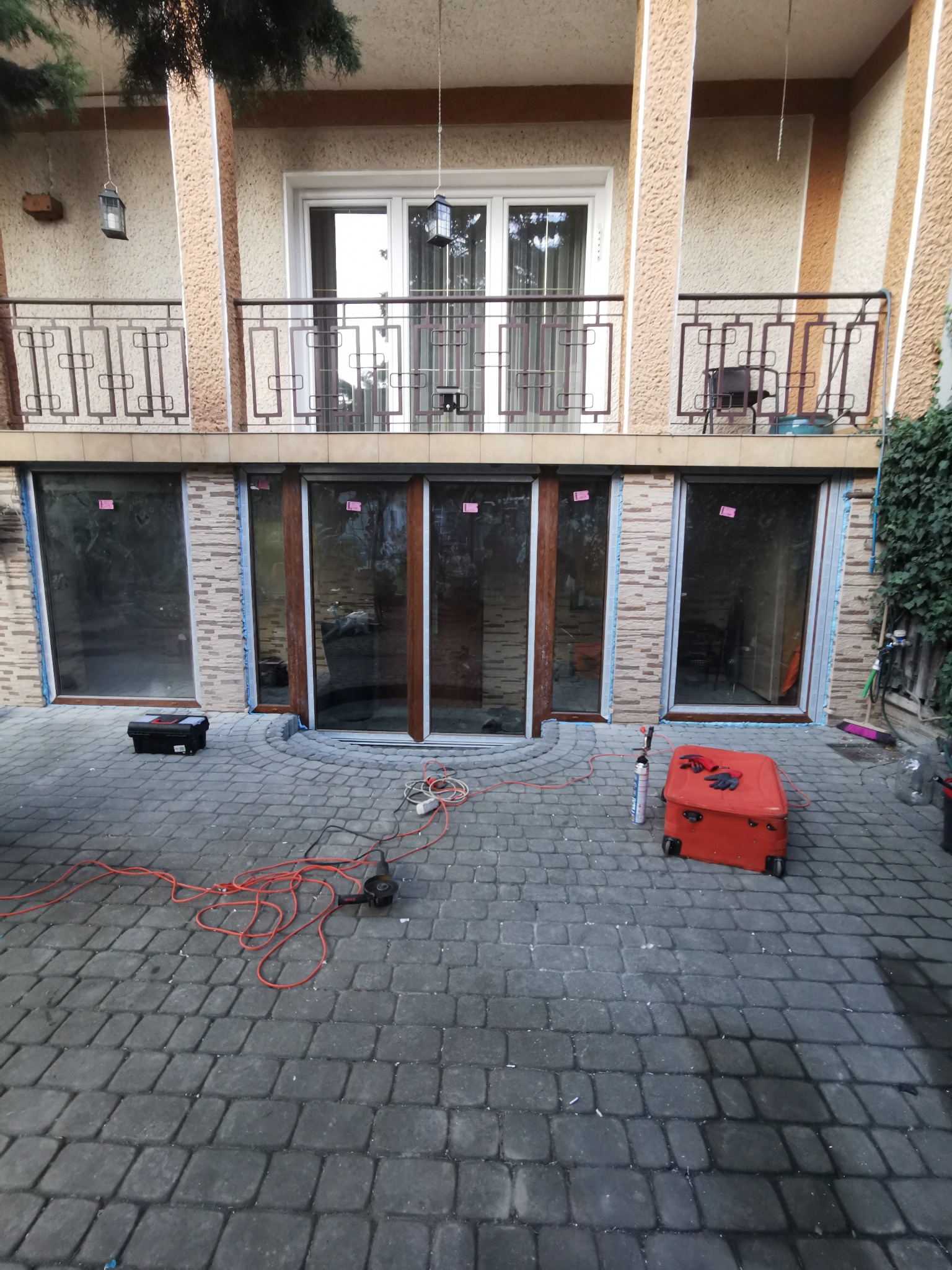 Montaż okien w budynku dwukondygnacyjnym, widoczne narzędzia, czerwona walizka na kółkach i pianka montażowa na brukowanym podłożu.