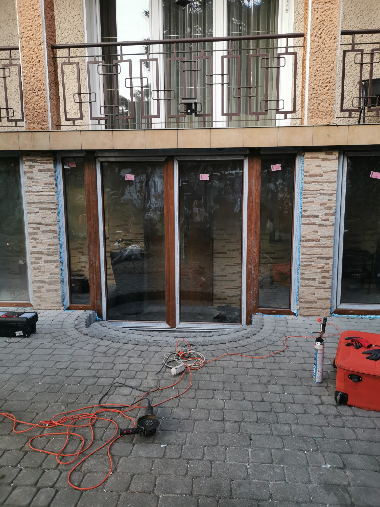 Montaż okien z brązowymi ramami w budynku z balkonem, widoczne narzędzia i materiały budowlane na brukowanym podłożu.