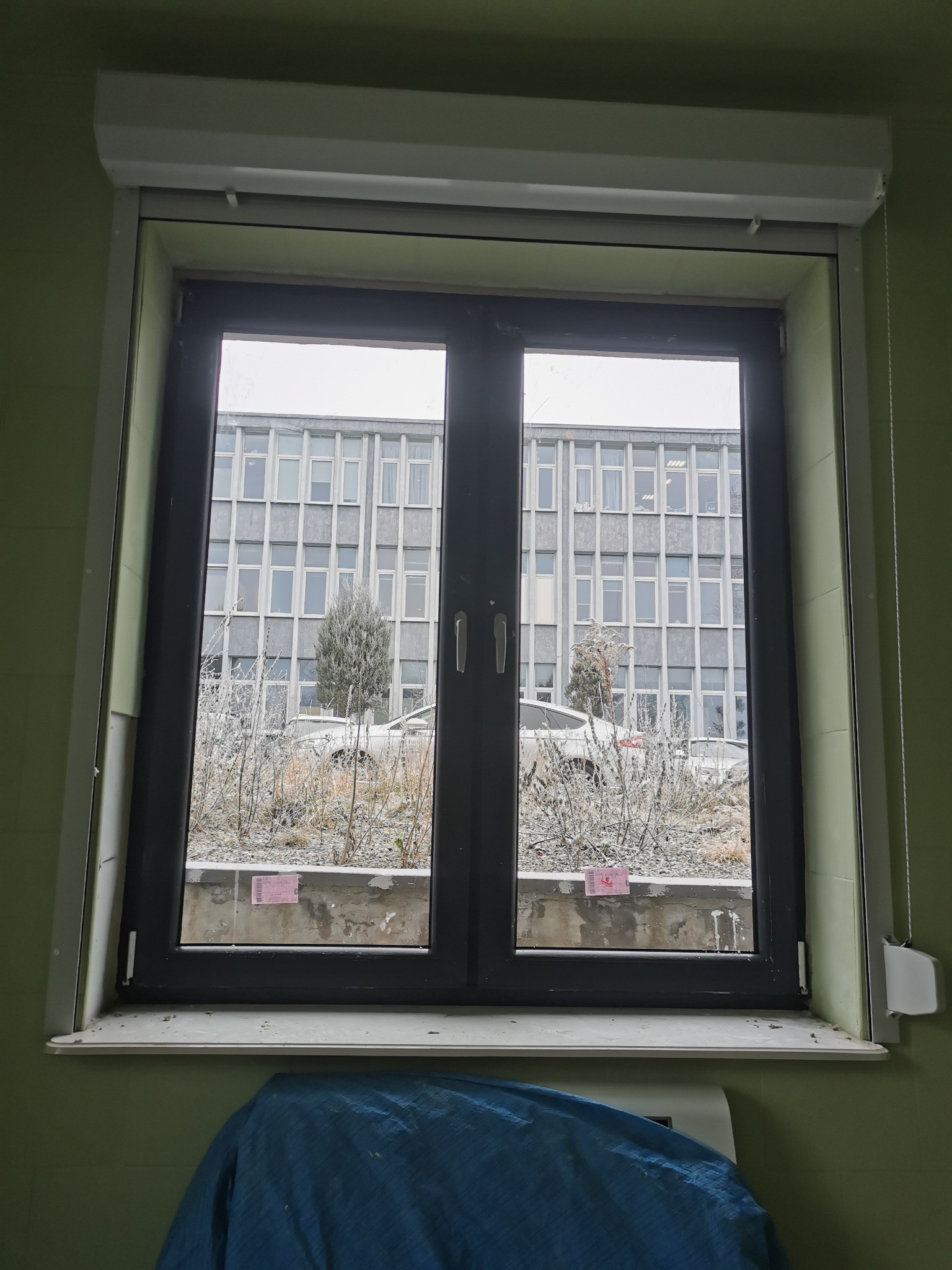 Dwuskrzydłowe okno PCV w kolorze antracytowym, z roletą zwiniętą u góry, widok na szary budynek z licznymi oknami i oszronioną roślinność na zewnątrz, parapet z naklejkami i fragmentem niebieskiej...