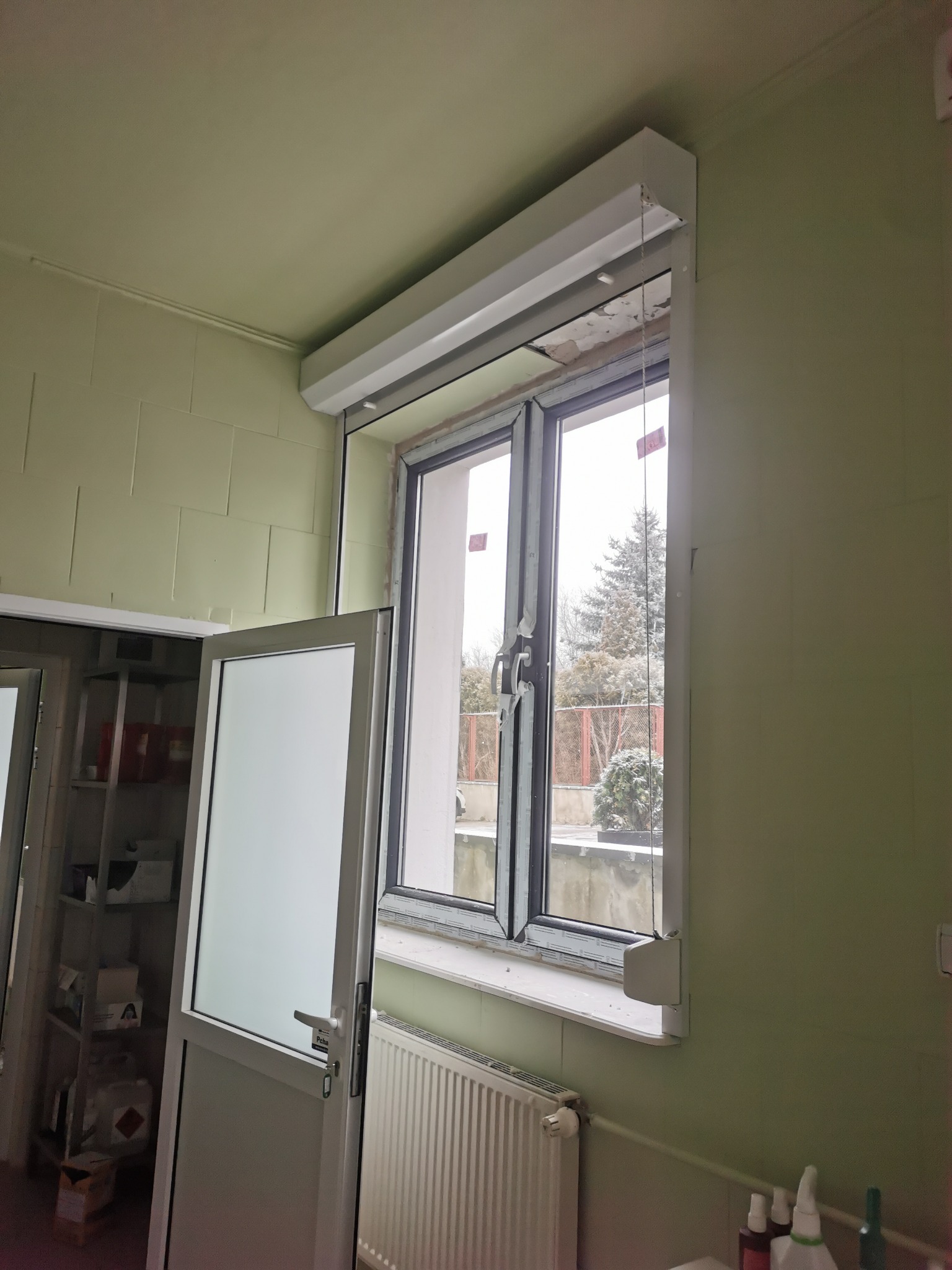 Okno z roletą zewnętrzną, widok z wnętrza pomieszczenia o zielonych ścianach, na zewnątrz widoczny zimowy krajobraz z drzewami i śniegiem.