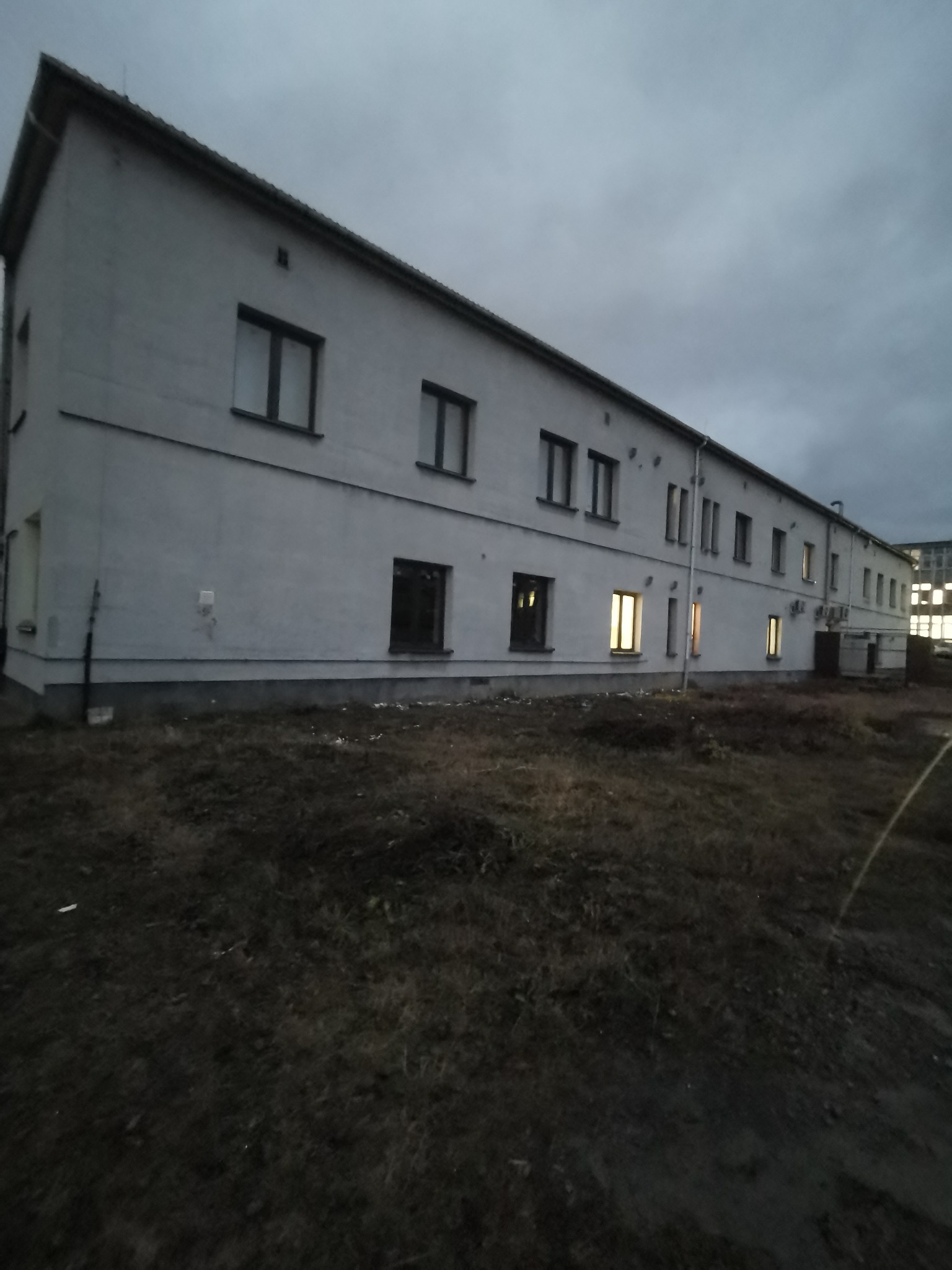 Długi, dwukondygnacyjny budynek z licznymi oknami, kilka z nich oświetlonych, sfotografowany z niskiej perspektywy podczas pochmurnego zmierzchu. Ziemia przed budynkiem porośnięta niską, ciemną...