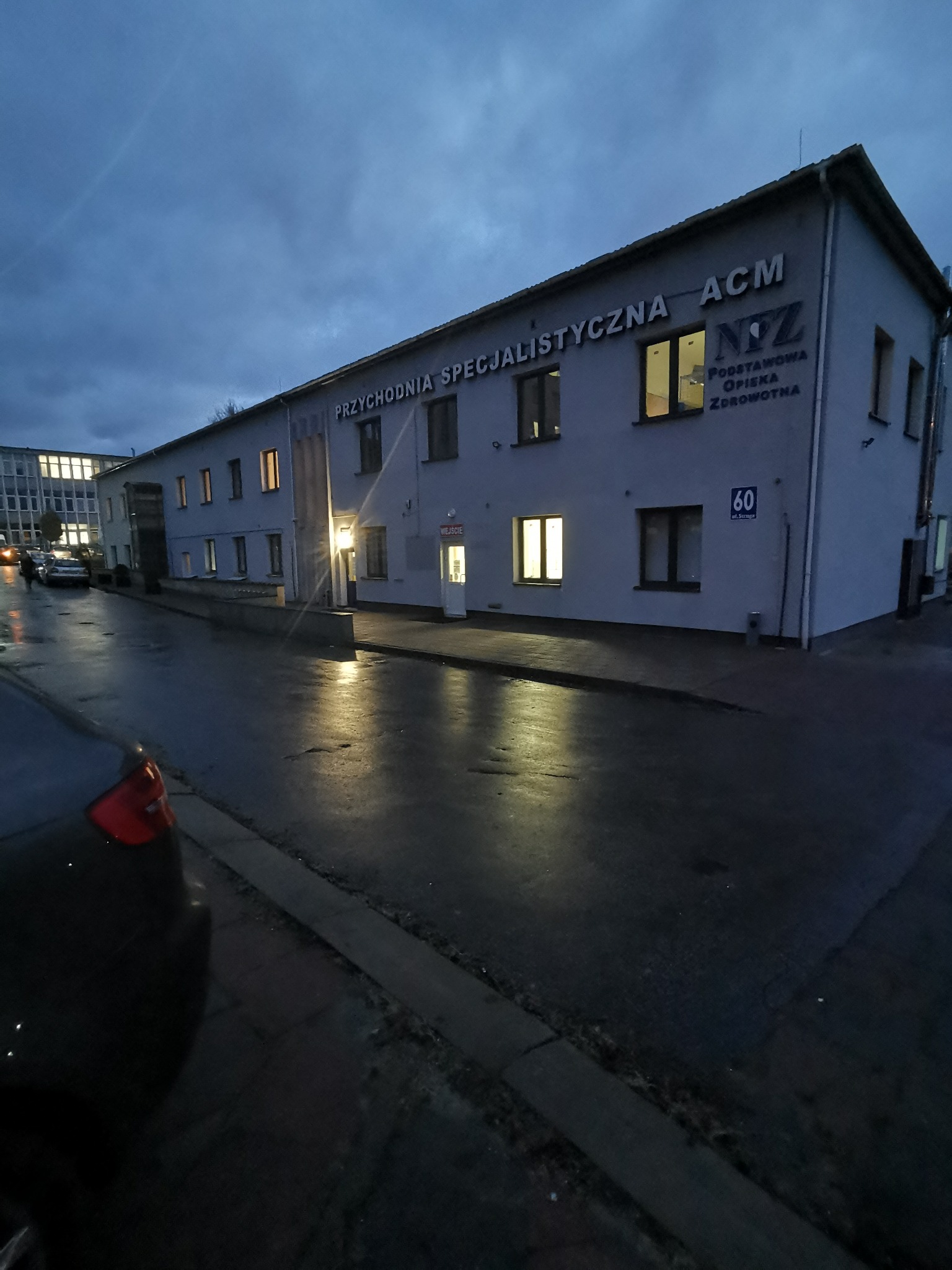 Budynek przychodni specjalistycznej ACM w pochmurny wieczór, mokra ulica odbija światła. Widoczne okna z włączonym światłem i tabliczka z numerem 60.