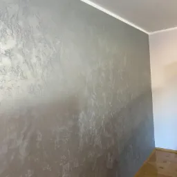 Ściana z dekoracyjną, srebrzystą fakturą, widoczna listwa przypodłogowa i fragment parkietu.